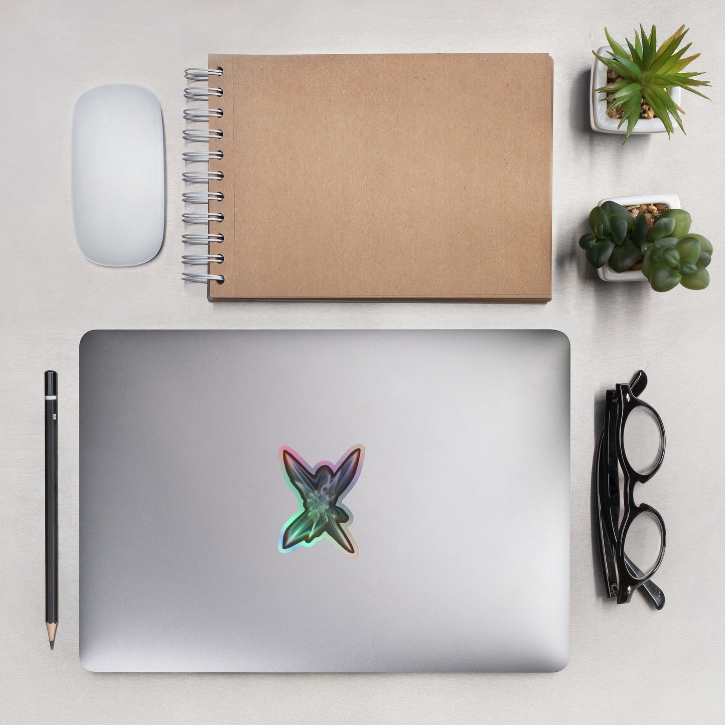 The Light Within Holographic sticker on a laptop with other items next to it to show scale including eyeglasses, plants, a pencil, mouse, and notebook.