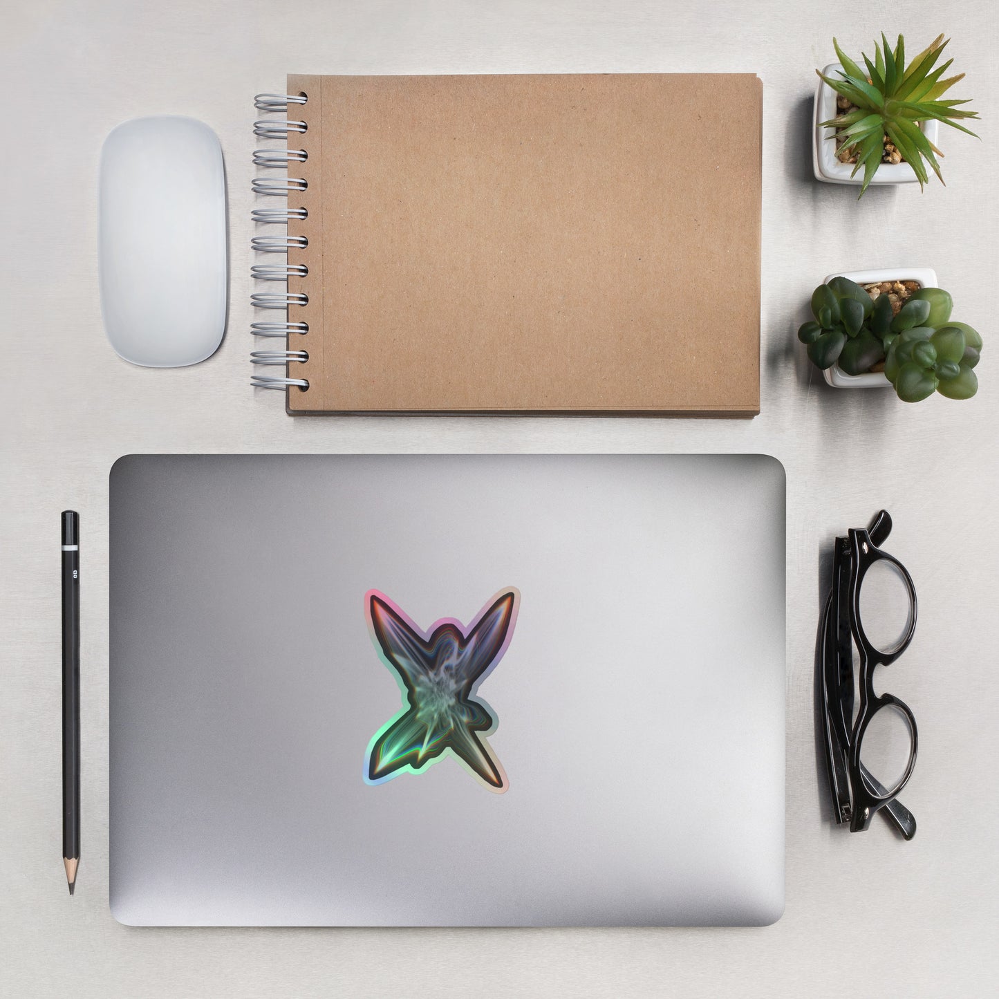 The Light Within Holographic sticker on a laptop with other items next to it to show scale including eyeglasses, plants, a pencil, mouse, and notebook.