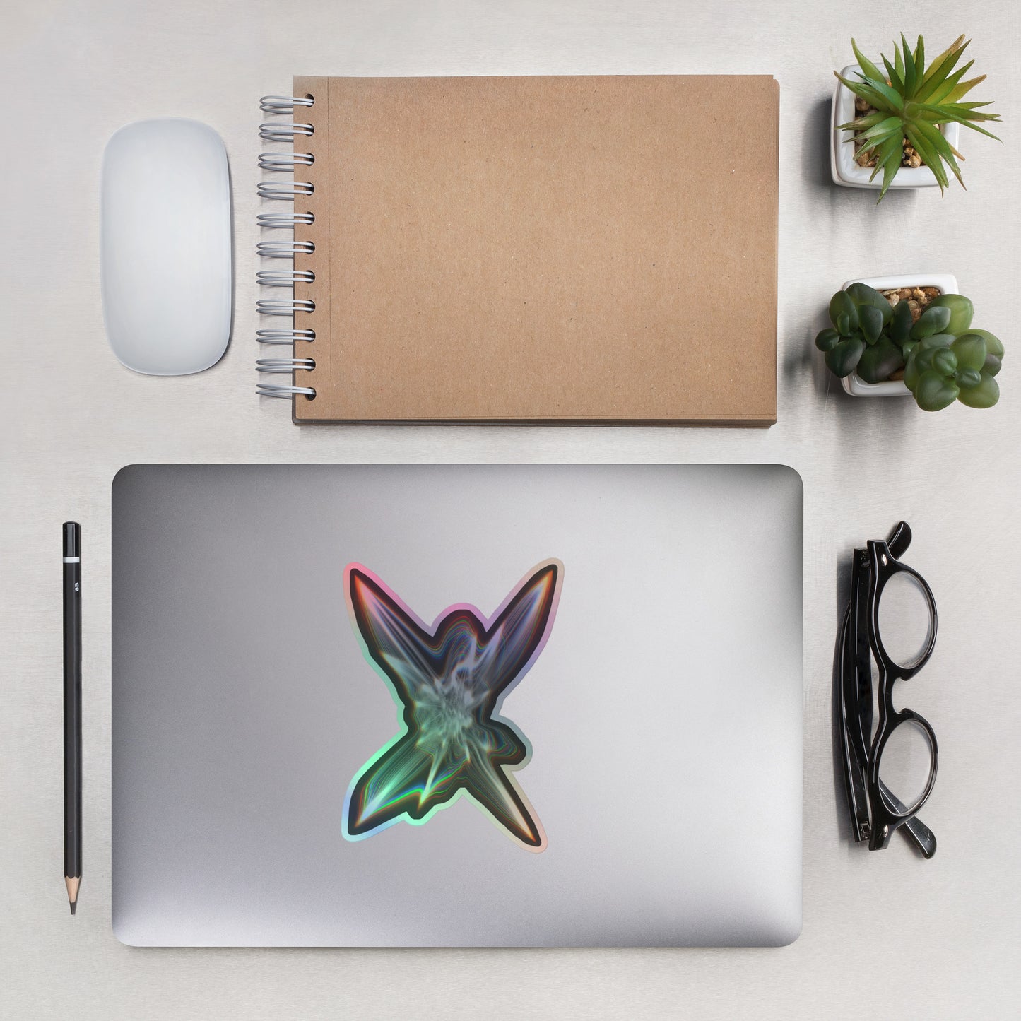 The Light Within Holographic sticker on a laptop with other items next to it to show scale including eyeglasses, plants, a pencil, mouse, and notebook.