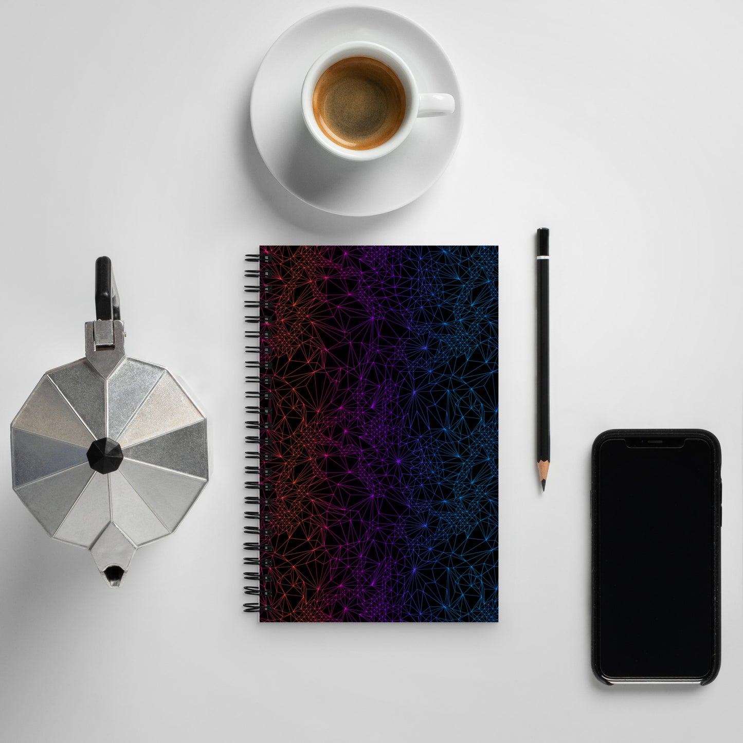 The Spiral notebook with Dark Gradient Tri-Mesh Geometric Pattern sitting on a table alongside some objects including a pencil, phone, espresso cup, and espresso pot.